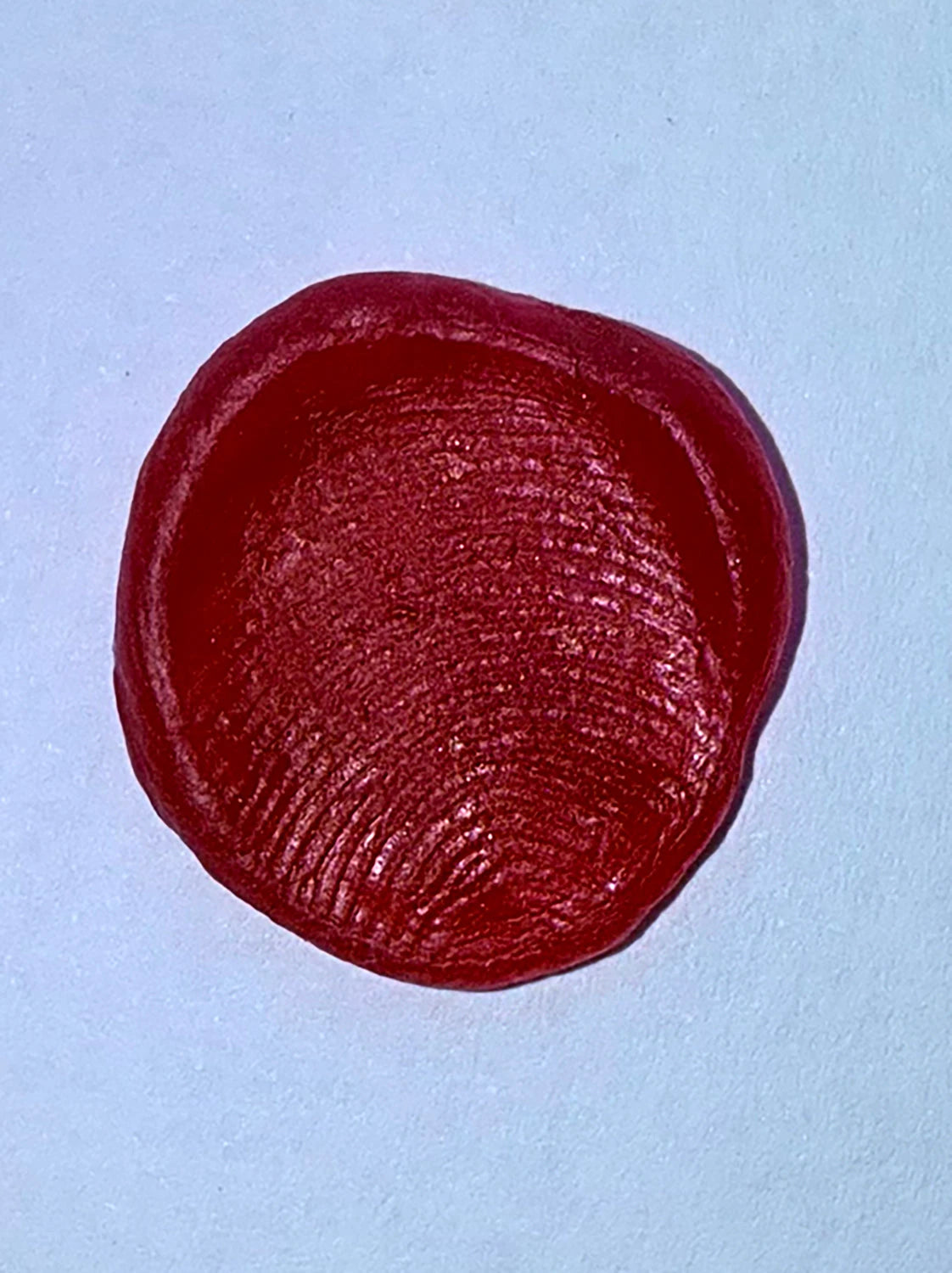 Close-up of a circular red wax impression, capturing fine texture details and fingerprint patterns. The wax has a smooth, slightly domed surface with subtle grooves, set against a light background.